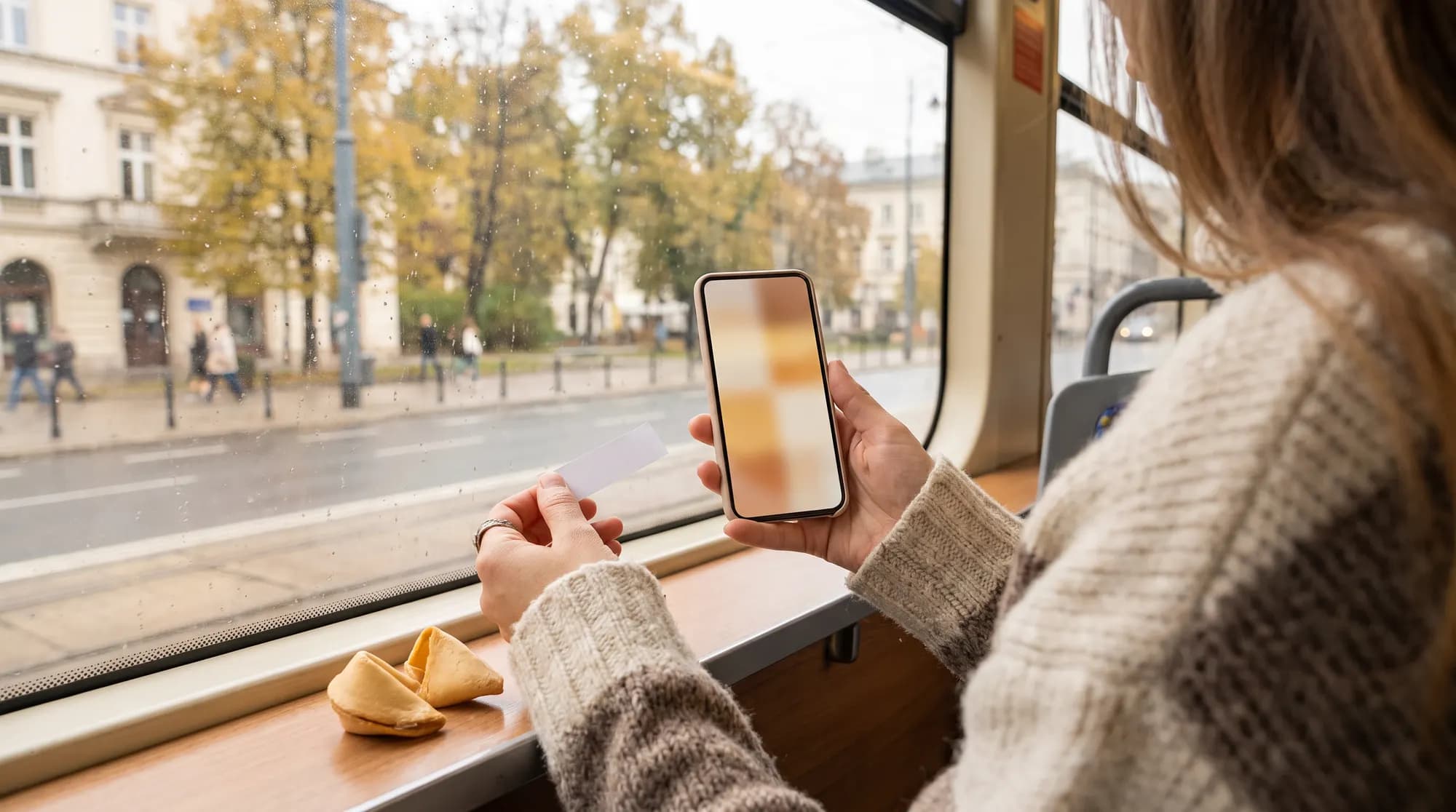 Pasażerka warszawskiego tramwaju trzyma smartfon i pustą karteczkę z ciasteczka z wróżbą, na parapecie leży przełamane ciasteczko, za oknem jesienna ulica